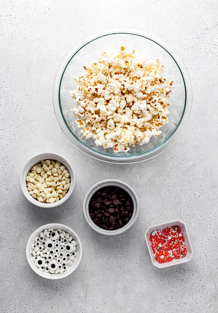 Overhead image of bowls of popped popcorn, white chocolate chips, dark chocolate chips, candy eyes, and orange and white sprinkles.