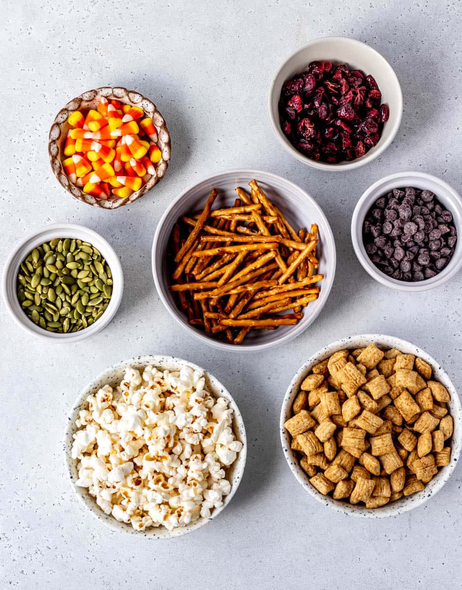 Bowls of cereal, popcorn, pretzels, pumpkin seeds, cranberries, dark chocolate chips, & candy corn for Halloween trail mix recipe