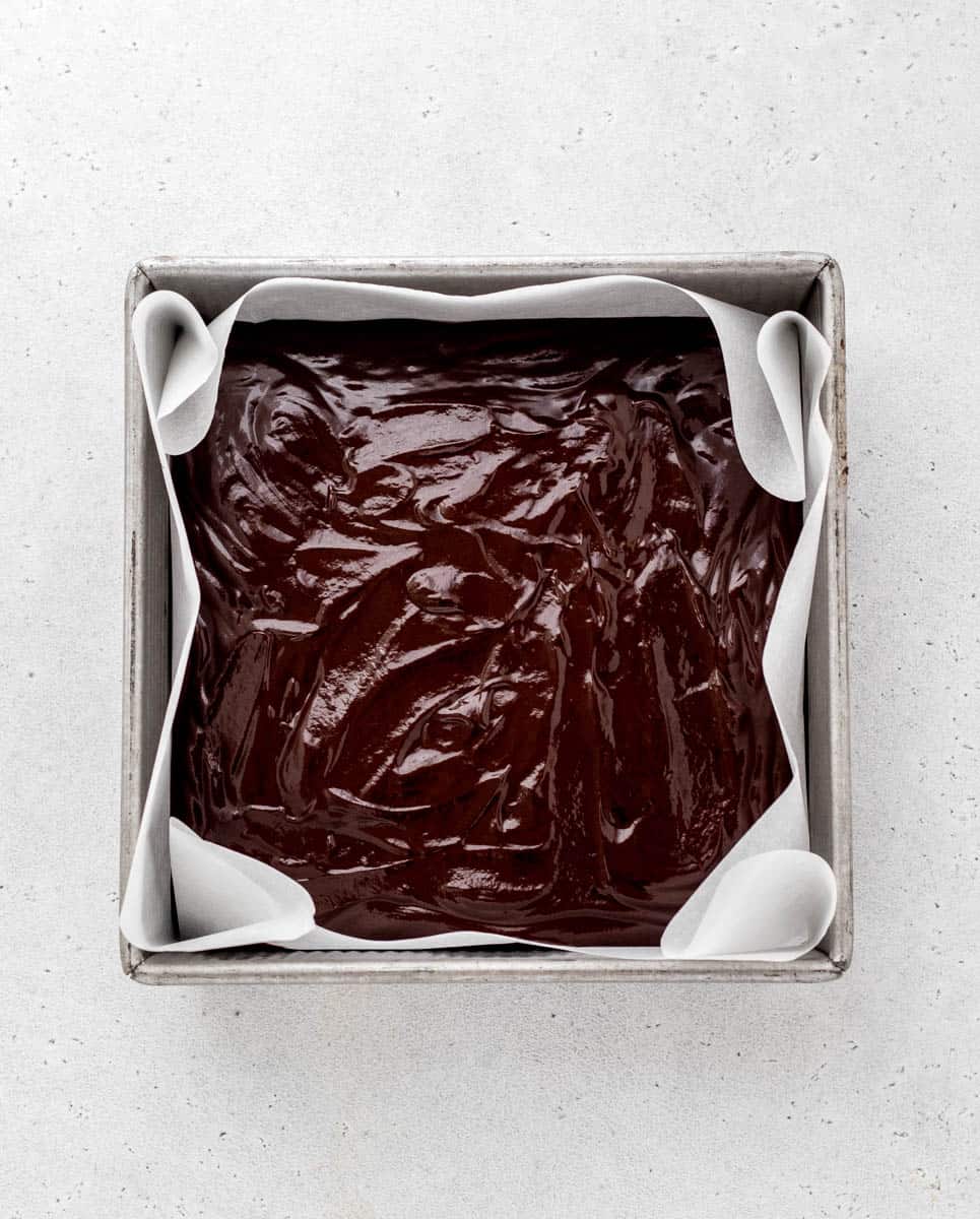 An 8x8 pan lined with parchment paper, filled with 3 ingredient peppermint fudge ready to be refrigerated.
