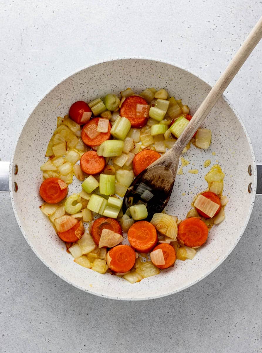 Carrots, onions, celery, and garlic sautéeing in a saucepan being stirred with a wooden spoon.