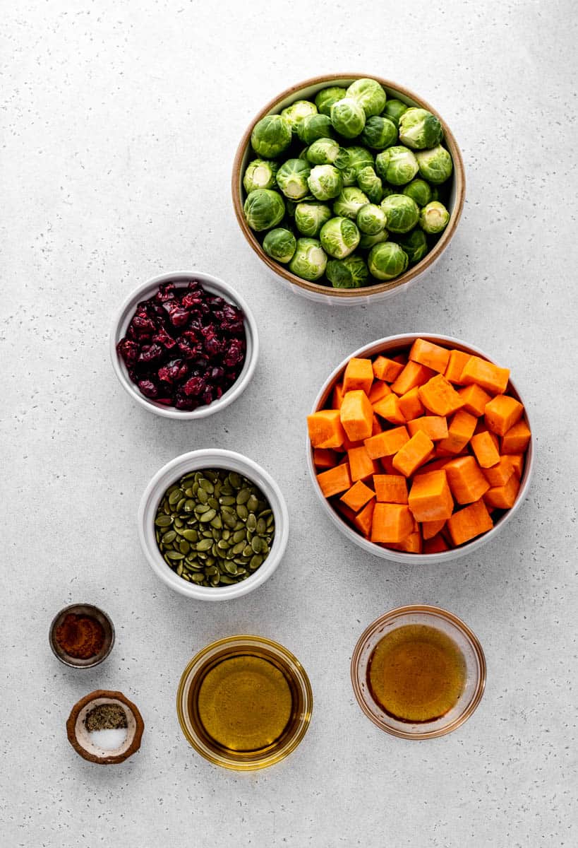 Separate bowls of brussel sprouts, cubed sweet potato, dried cranberries, unsalted pumpkin seeds, salt and pepper, olive oil, maple syrup, and cinnamon.