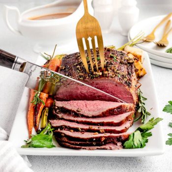 A fork holding Christmas beef roast while a sharp knife carves the roast.