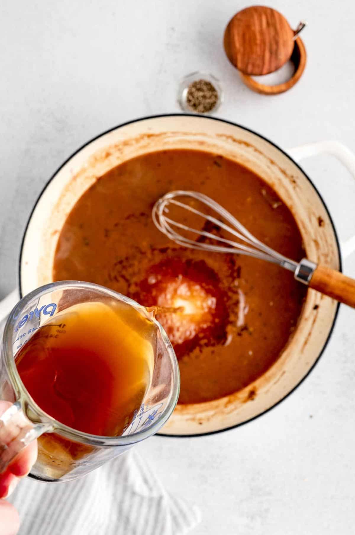 Beef broth being poured into the pot of homemade gravy, a whisk is ready to mix it.