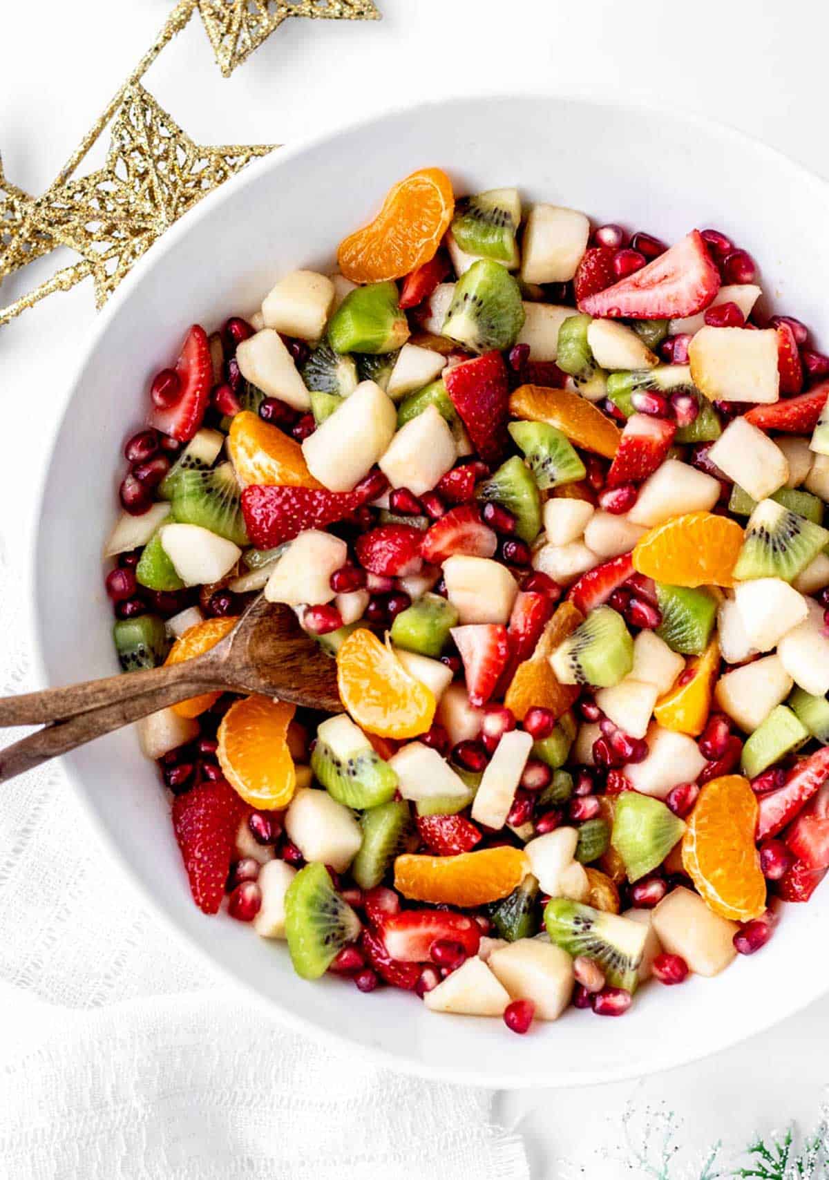 A large white serving bowl of mixed winter fruit salad, with a wooden serving spoon.