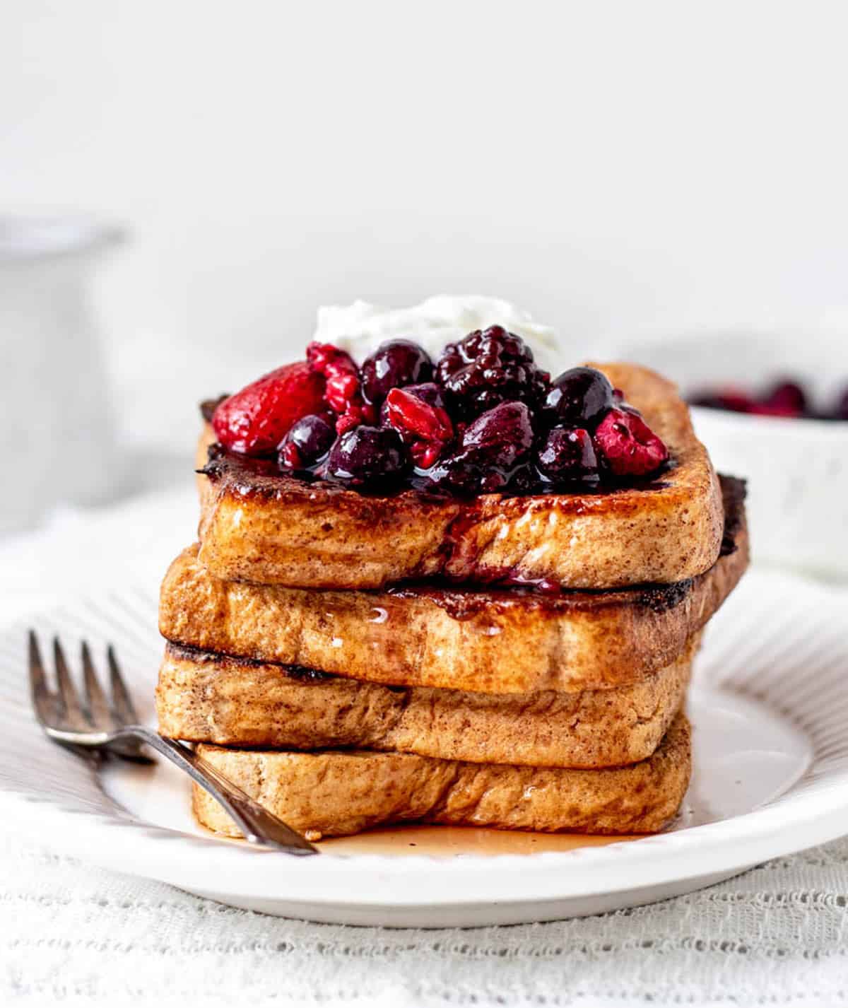 4 slices of high protein French toast stacked on a white plate, topped with mixed berries, Greek yogurt and maple syrup.