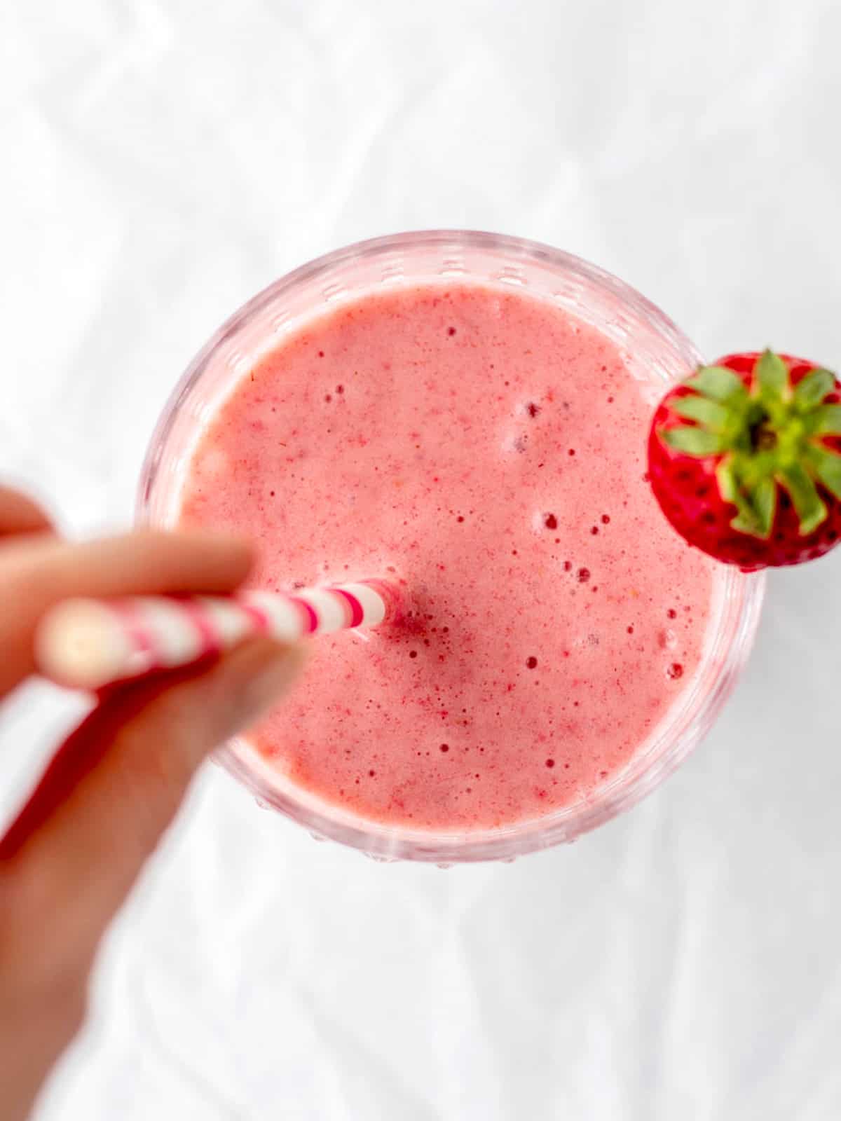 A hand holding the straw in a glass of healthy strawberry banana smoothie.