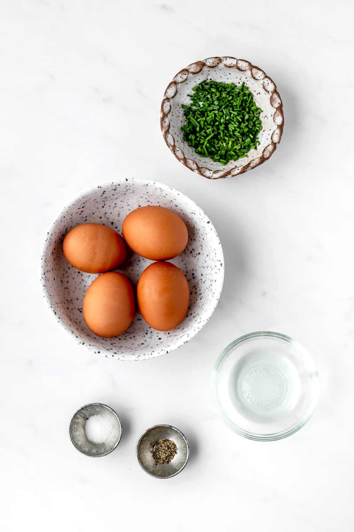 The ingredients for creamy scrambled eggs; including 4 eggs, salt and pepper, and olive oil, plus chives for a garnish.