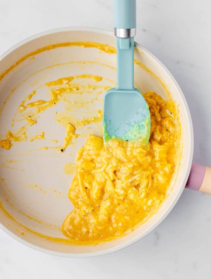 A non stick frying pan with soft scrambled eggs being pushed with a rubber spatula.