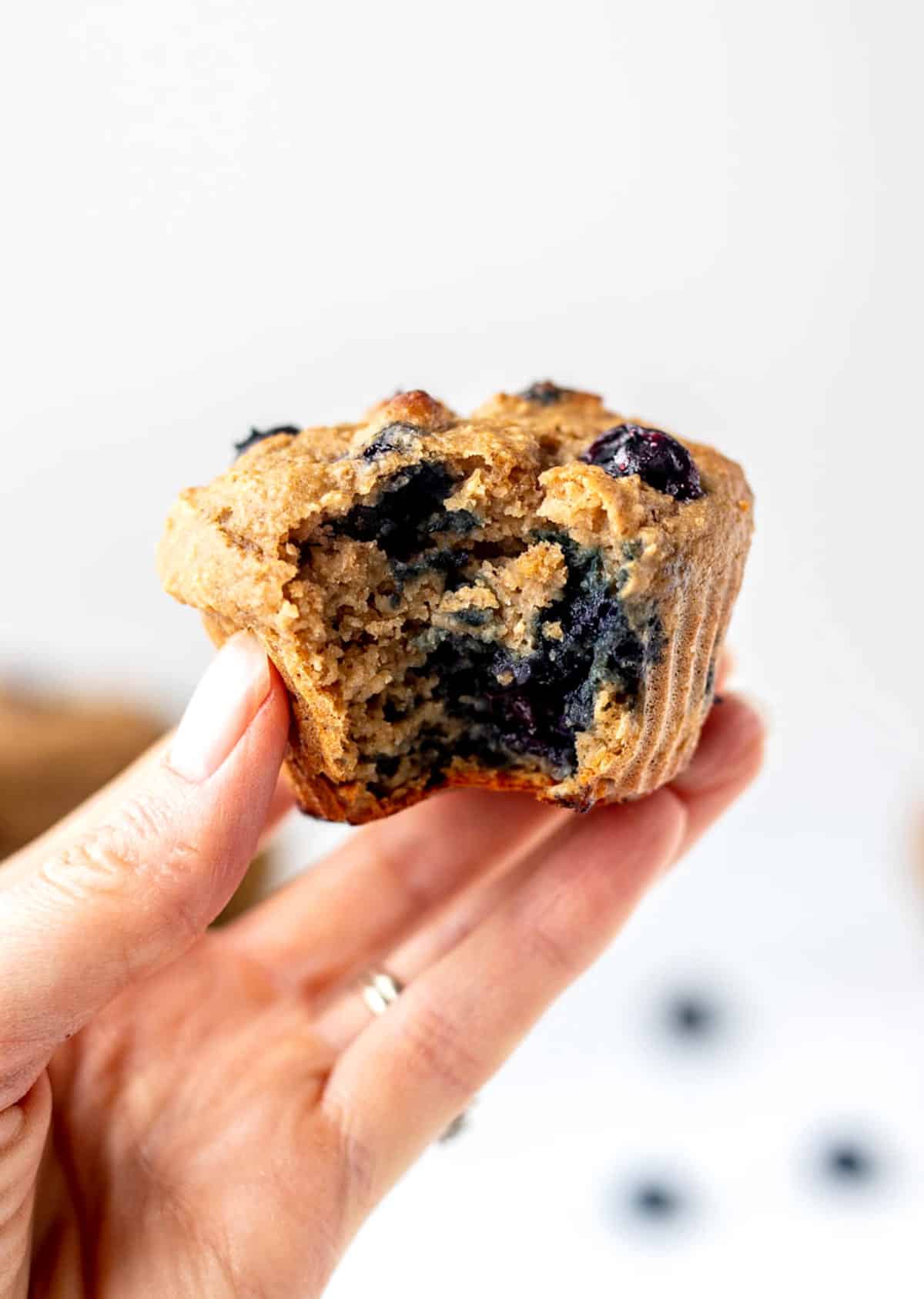 A hand holding up a blueberry protein muffin with a bite taken out of it.