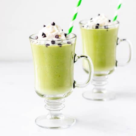 Two shamrock protein shakes in glasses topped with whipped cream and chocolate chips.