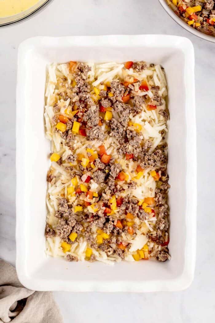 A layer of cooked ground sausage and veggies on top of hash browns in a baking dish.