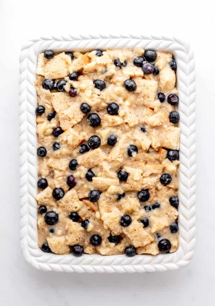 Egg mixture poured over the bread and blueberries in a white casserole dish.