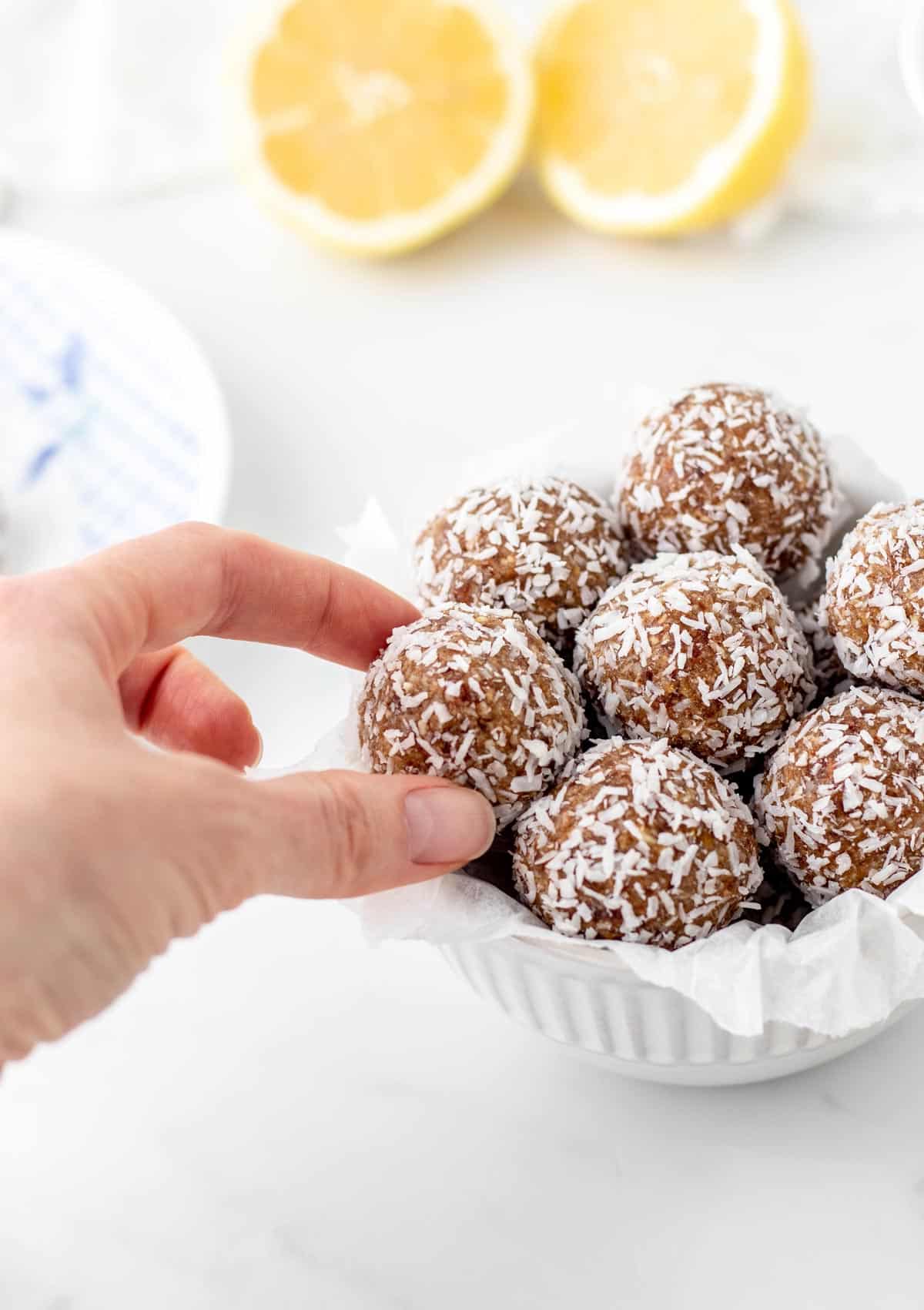 A hand grabbing one of the lemon bliss balls out of a small white bowl.