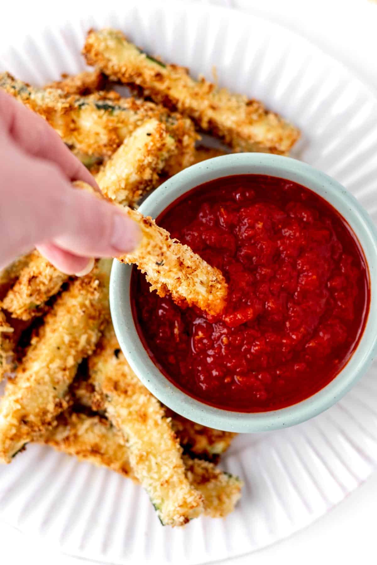 A hand dipping one of the parmesan zucchini fries in marinara sauce.