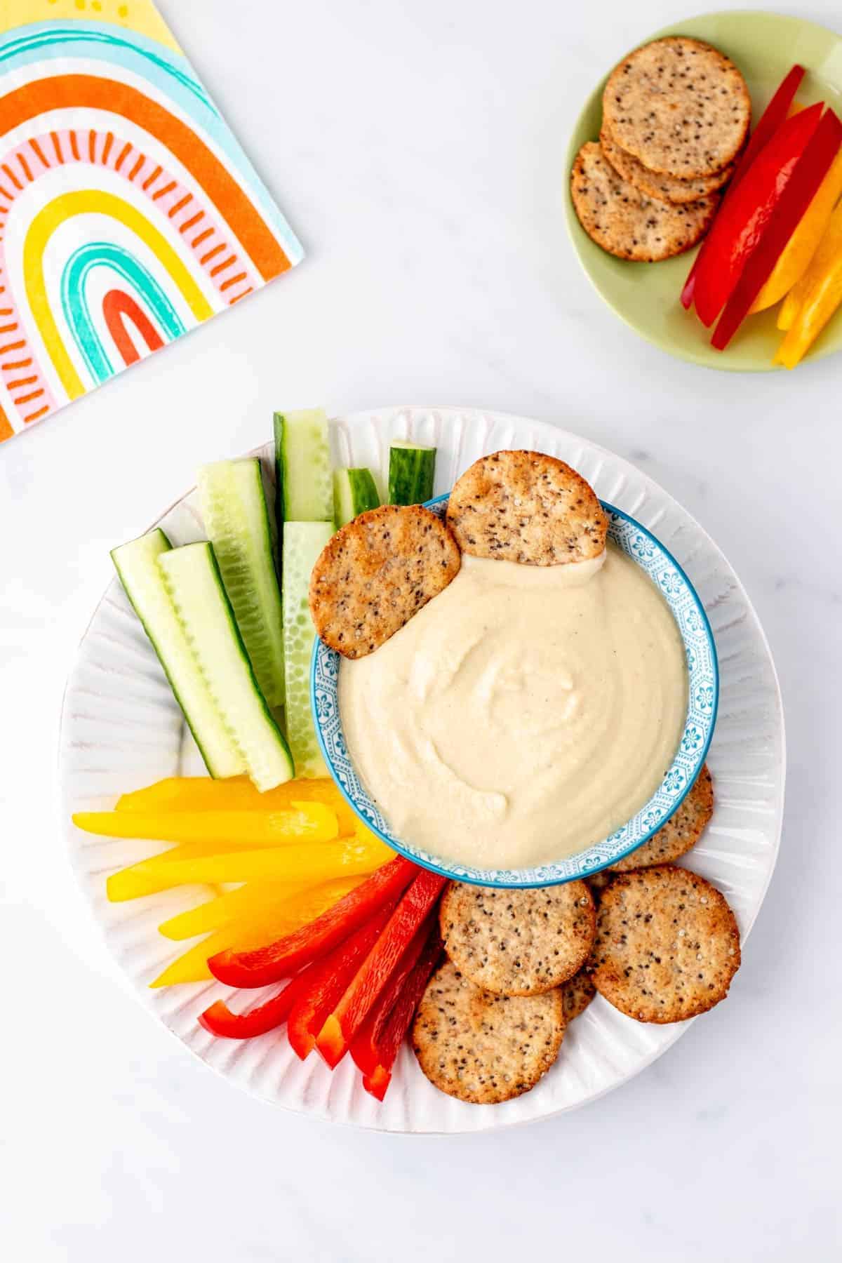 A plate of veggies and crackers with a bowl of high protein hummus.