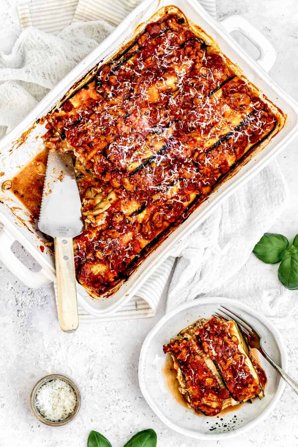 A slice of turkey vegetable lasagna on a plate next to a casserole dish and basil.