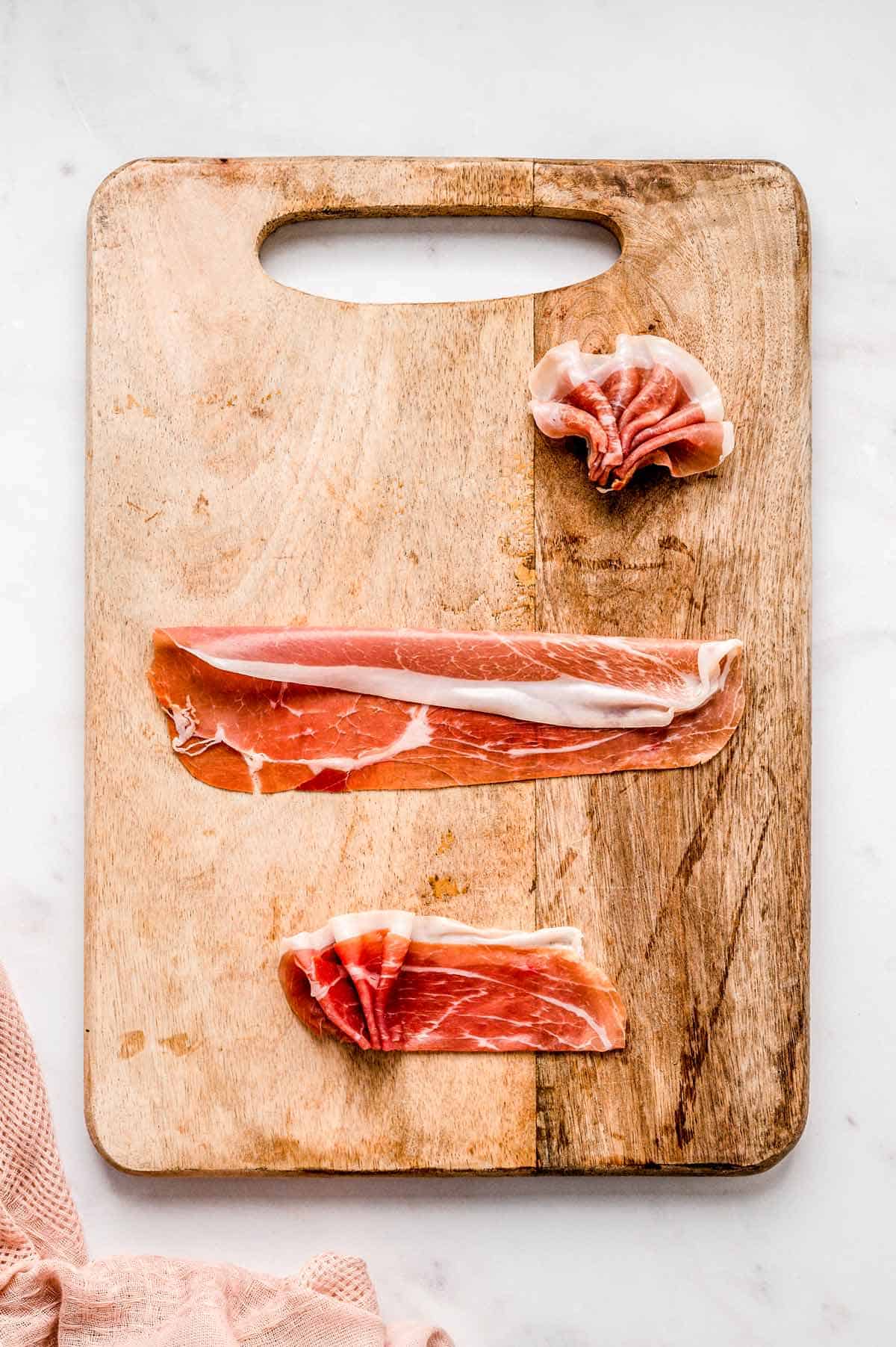 Thinly sliced meat on a wooden cutting board demonstrating how to make prosciutto fans.