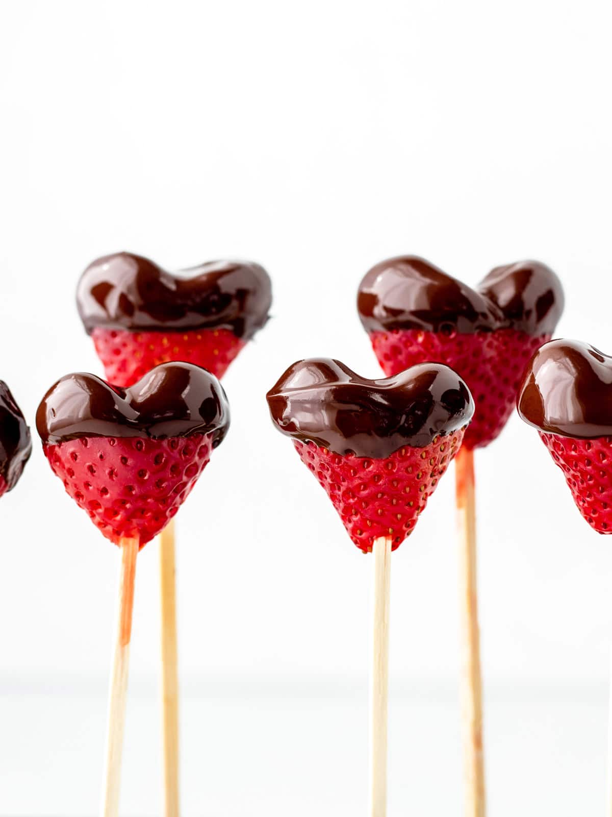 Chocolate covered strawberry hearts standing up on wooden skewers.
