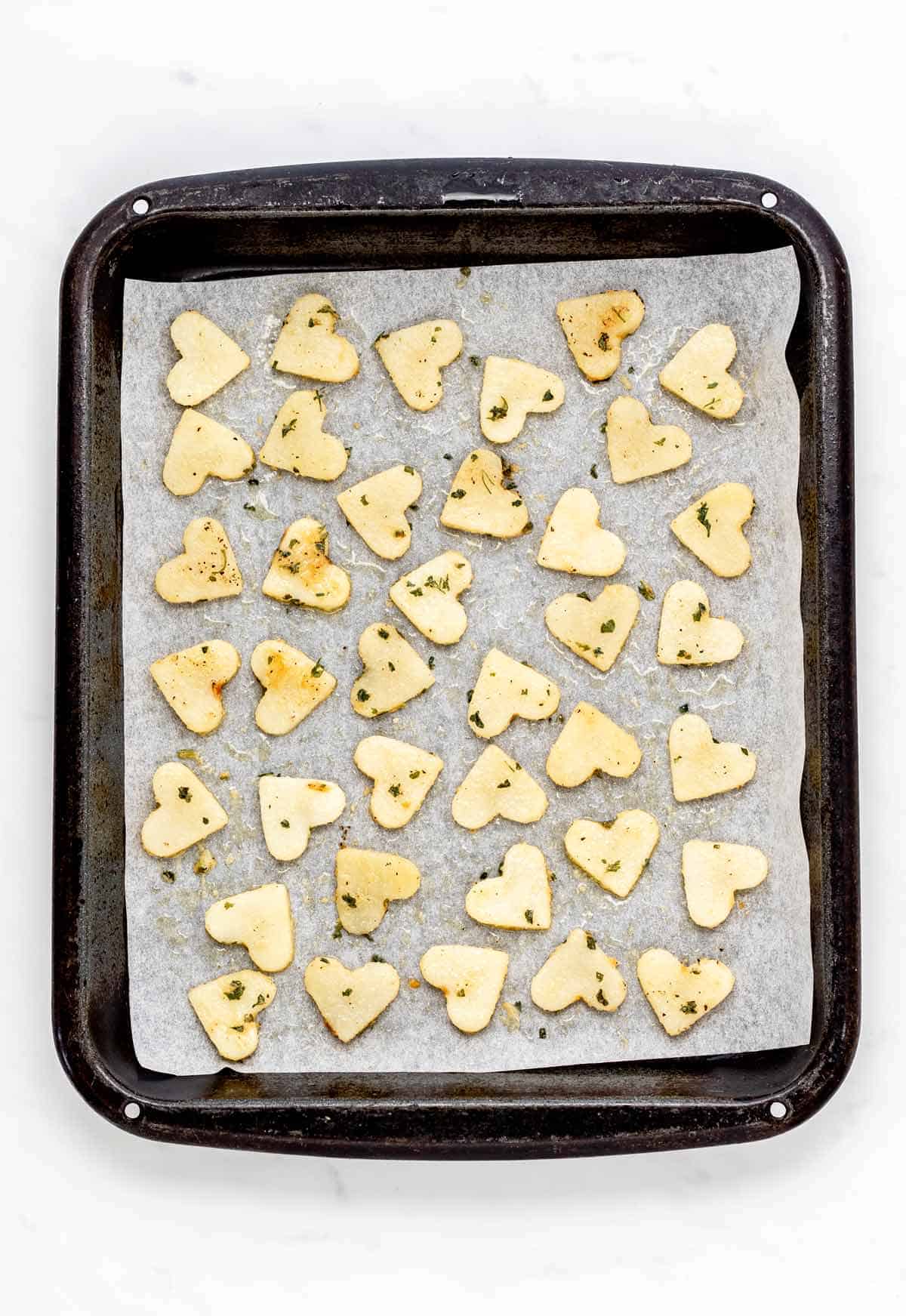 Heart shaped potato slices on a parchment paper lined baking sheet, prior to baking.