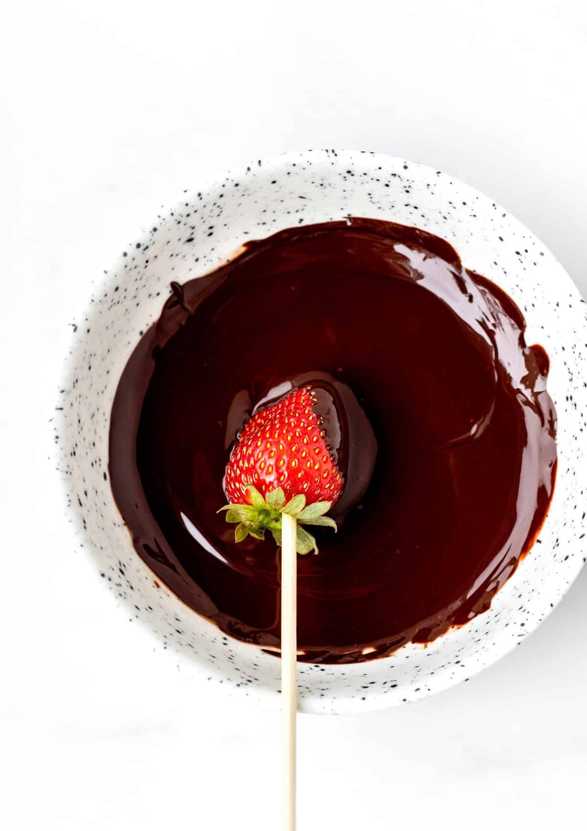 A strawberry on a stick being dipped into the melted chocolate.