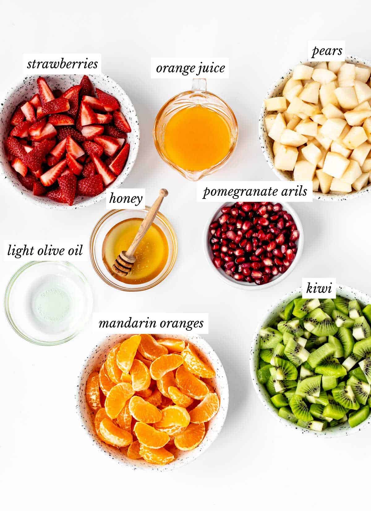 Labeled ingredients for Christmas fruit salad recipe, including strawberries, orange juice, pears, honey, pomegranate arils, mandarin oranges, and kiwi.