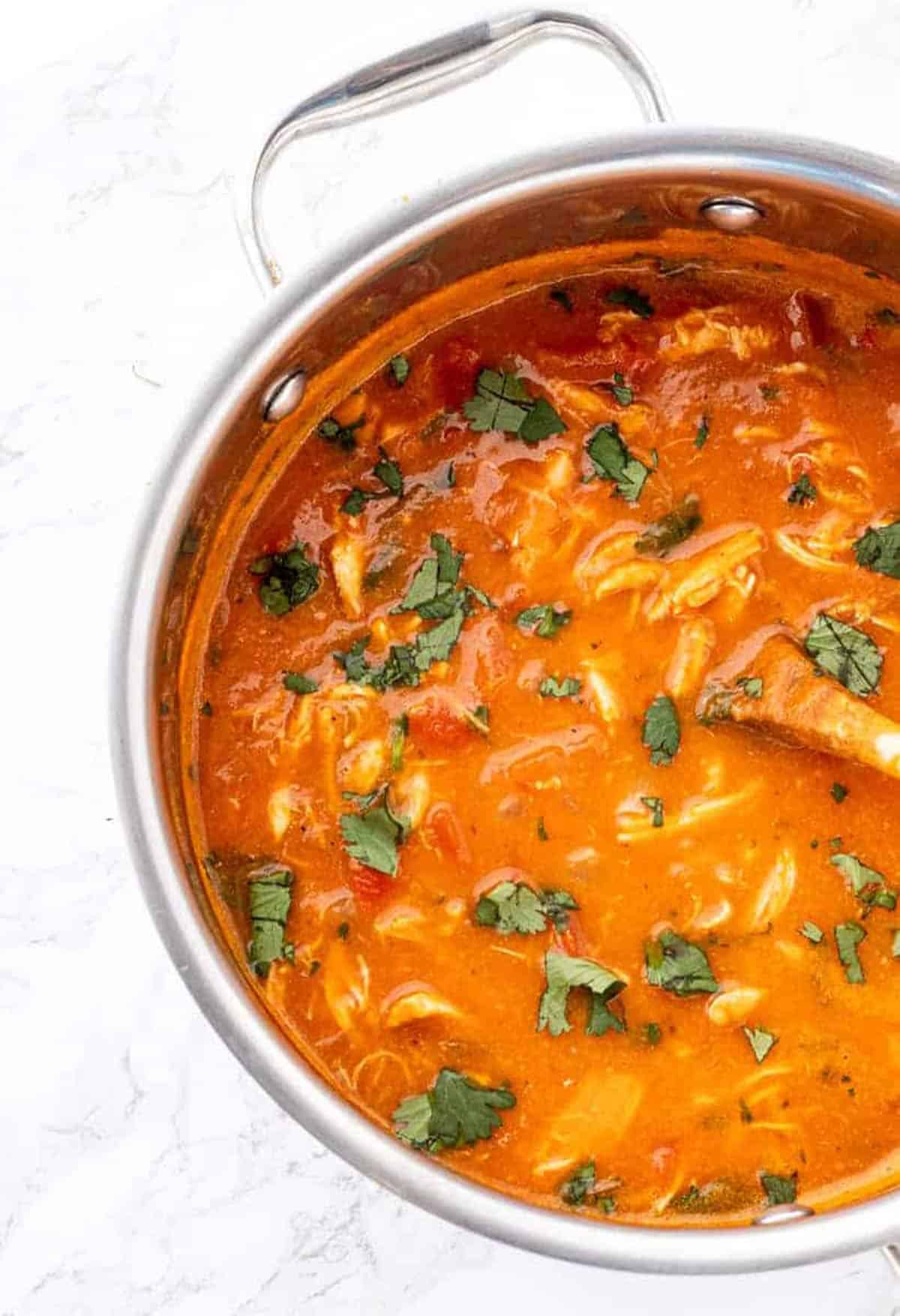 Large pot of pumpkin chicken soup with wooden spoon garnished with fresh cilantro on marble background.