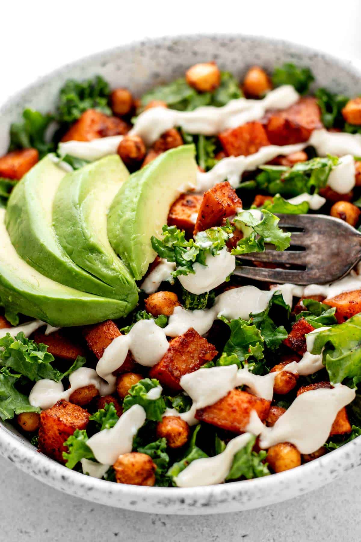 A fork digging into a bowl of sweet potato kale salad with roasted chickpeas and avocado.