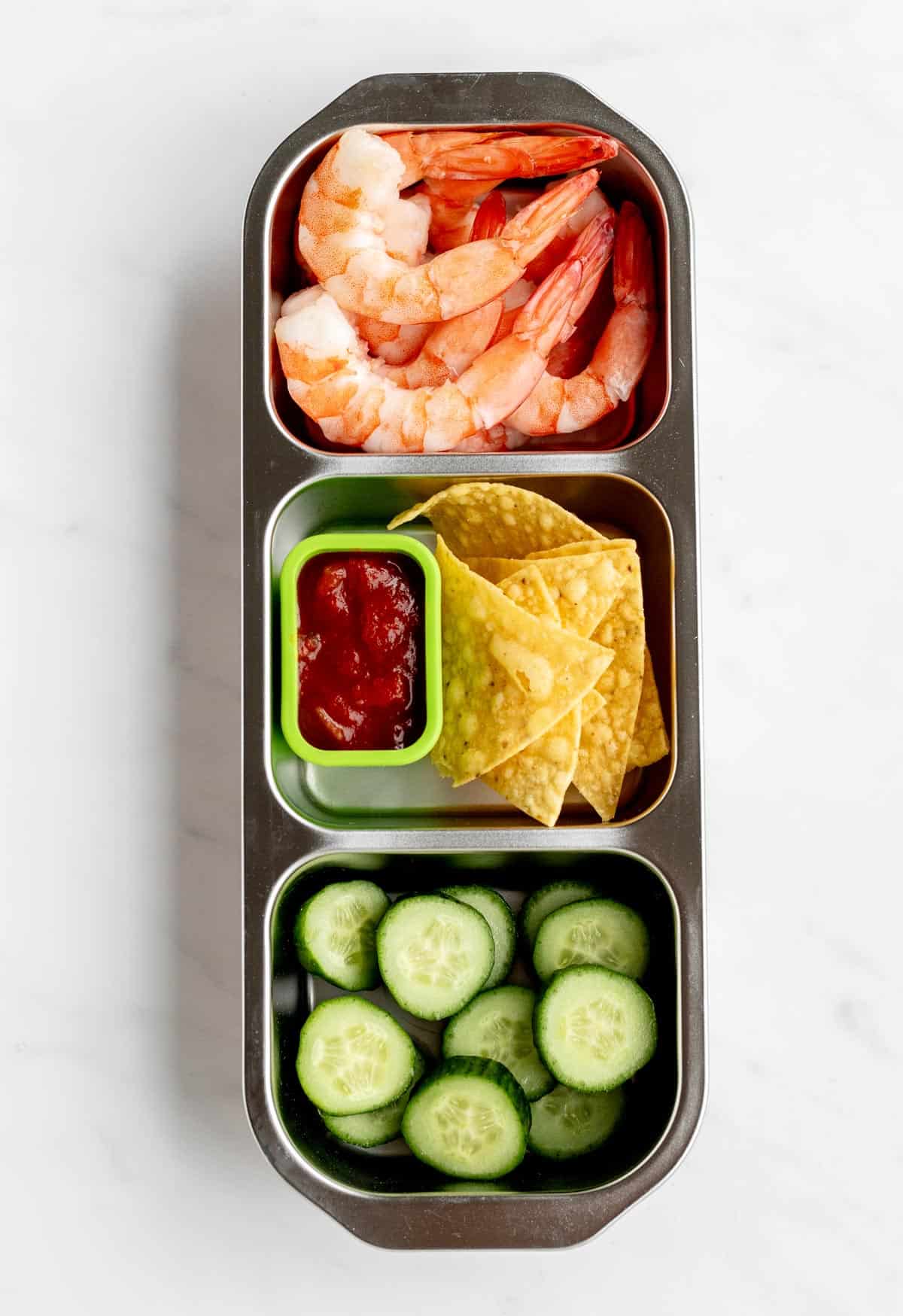 Bird's eye view of shrimp, tortilla chips, salsa, and cucumber slices in a three compartment snack box.