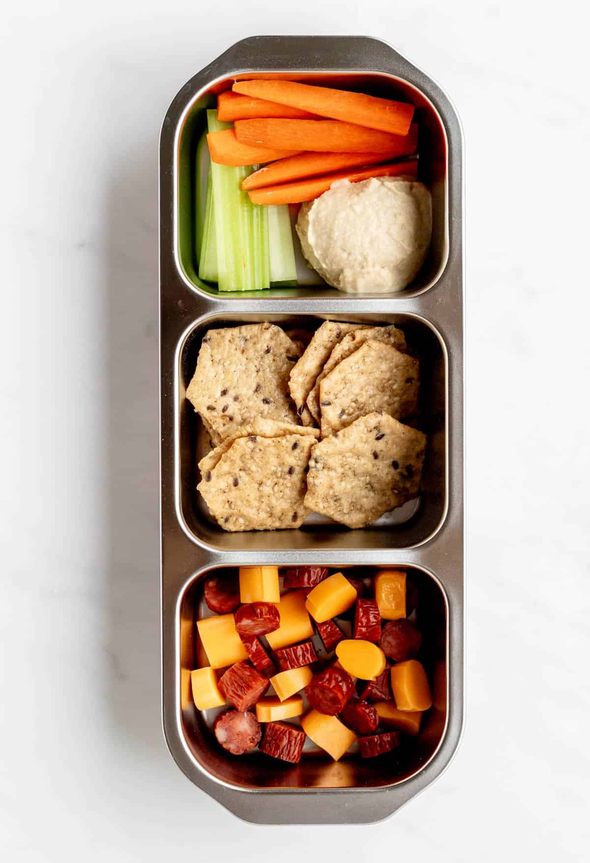 Bird's eye view of sliced turkey pepperette and cheese, crackers, veggies and hummus in a three compartment snack box.