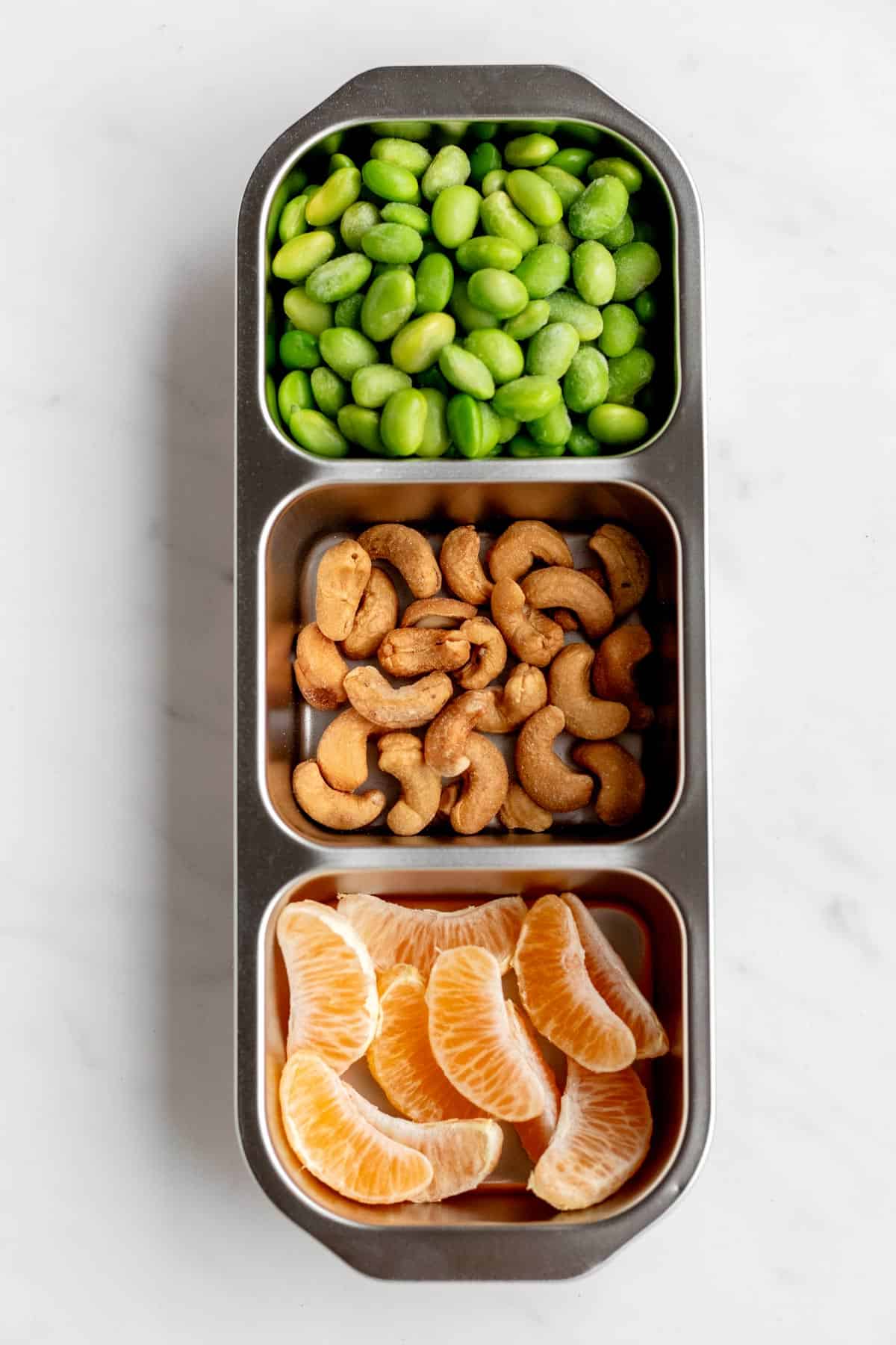 Bird's eye view of edamame, cashews and mandarin orange slices in a three compartment snack box.