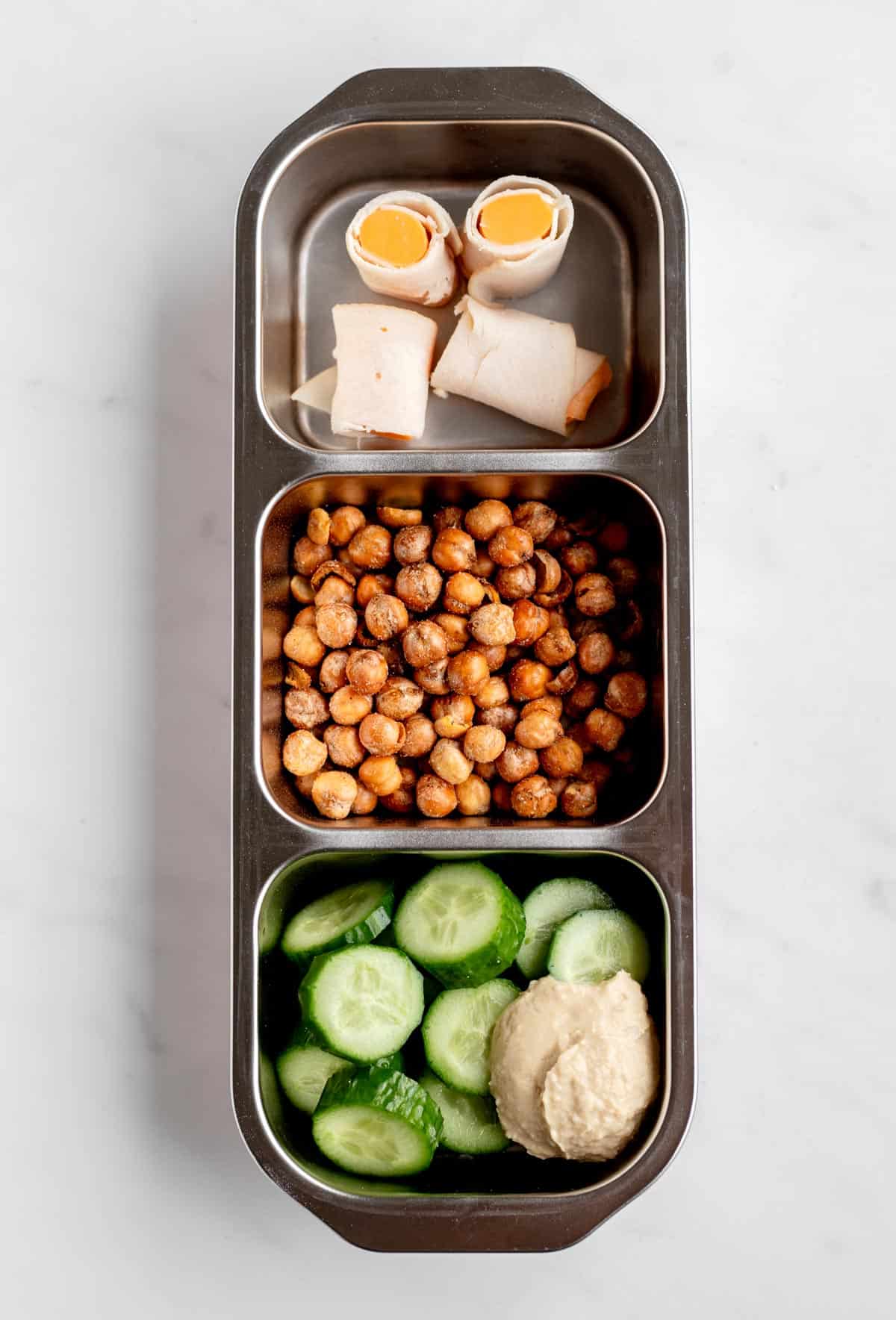 A snack box with turkey cheese roll ups, roasted chickpeas, cucumber slices and hummus.