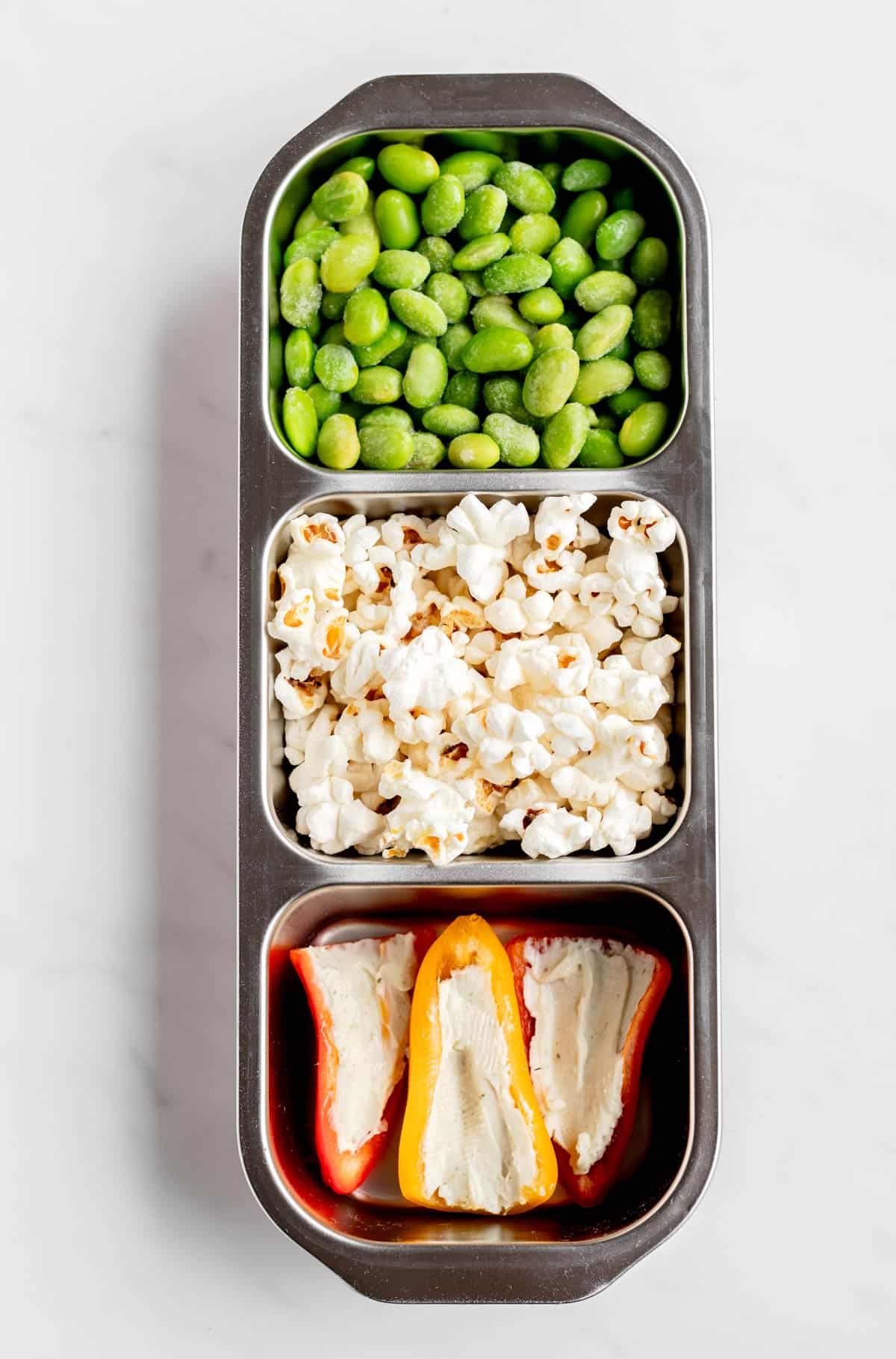 Edamame, popcorn and mini pepper halves filled with creamy cheese in a snack box.